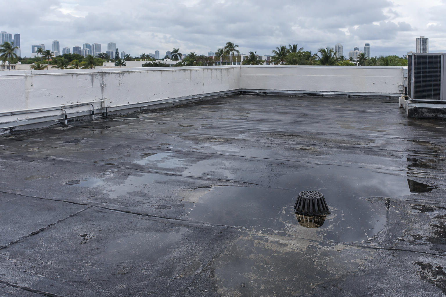 fotografía de techo comercial plano en Miami con charcos pequeños cerca de un drenaje, superficie asfáltica envejecida, parapeto blanco, luz nublada después de lluvia, sin personas, sin texto ni logotipos